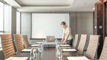 Un salón de conferencias con una larga mesa, varias sillas, una computadora portátil, botellas de agua y una pantalla de proyector.