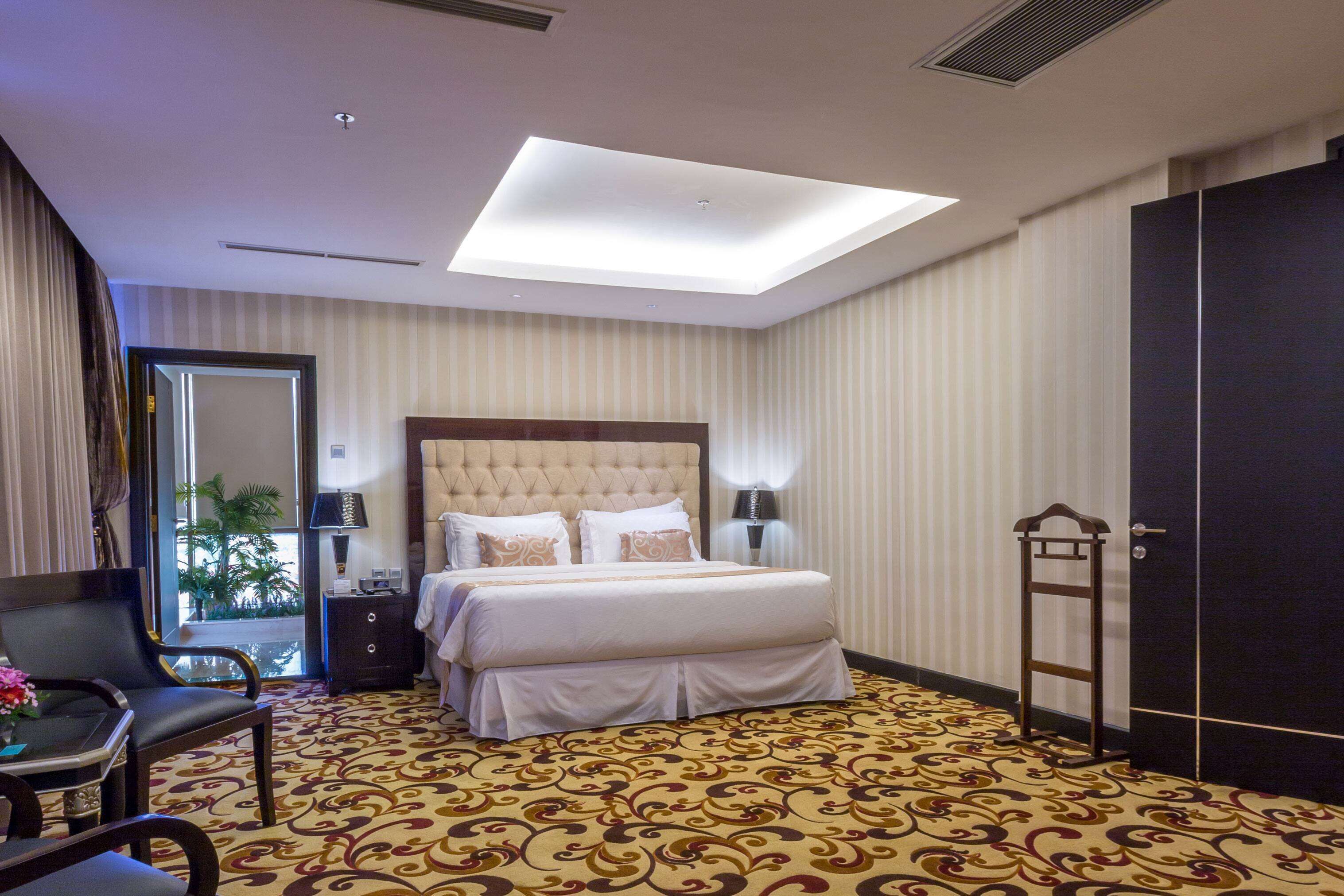 Habitación de hotel con una cama grande, un escritorio con silla, una mesita con flores, una mesita de noche, una lámpara, un armario y vistas al exterior a través de una ventana.