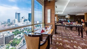 Habitación de hotel con un ventanal amplio que ofrece vistas a la ciudad, una zona de comedor con mesa para dos personas y una decoración interior moderna.