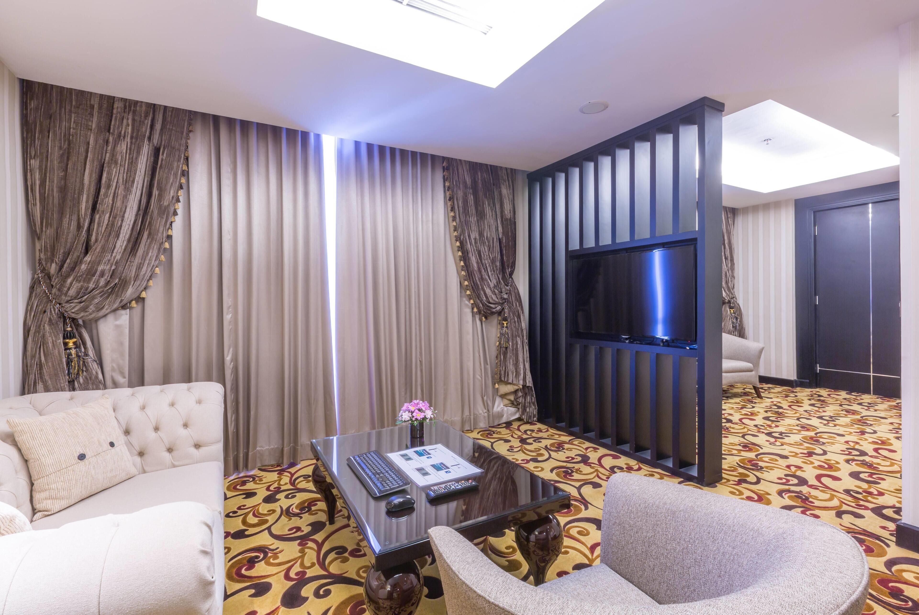 Habitación de hotel con sofá, sillón, una mesita de vidrio con un control remoto, televisión y alfombra estampada.