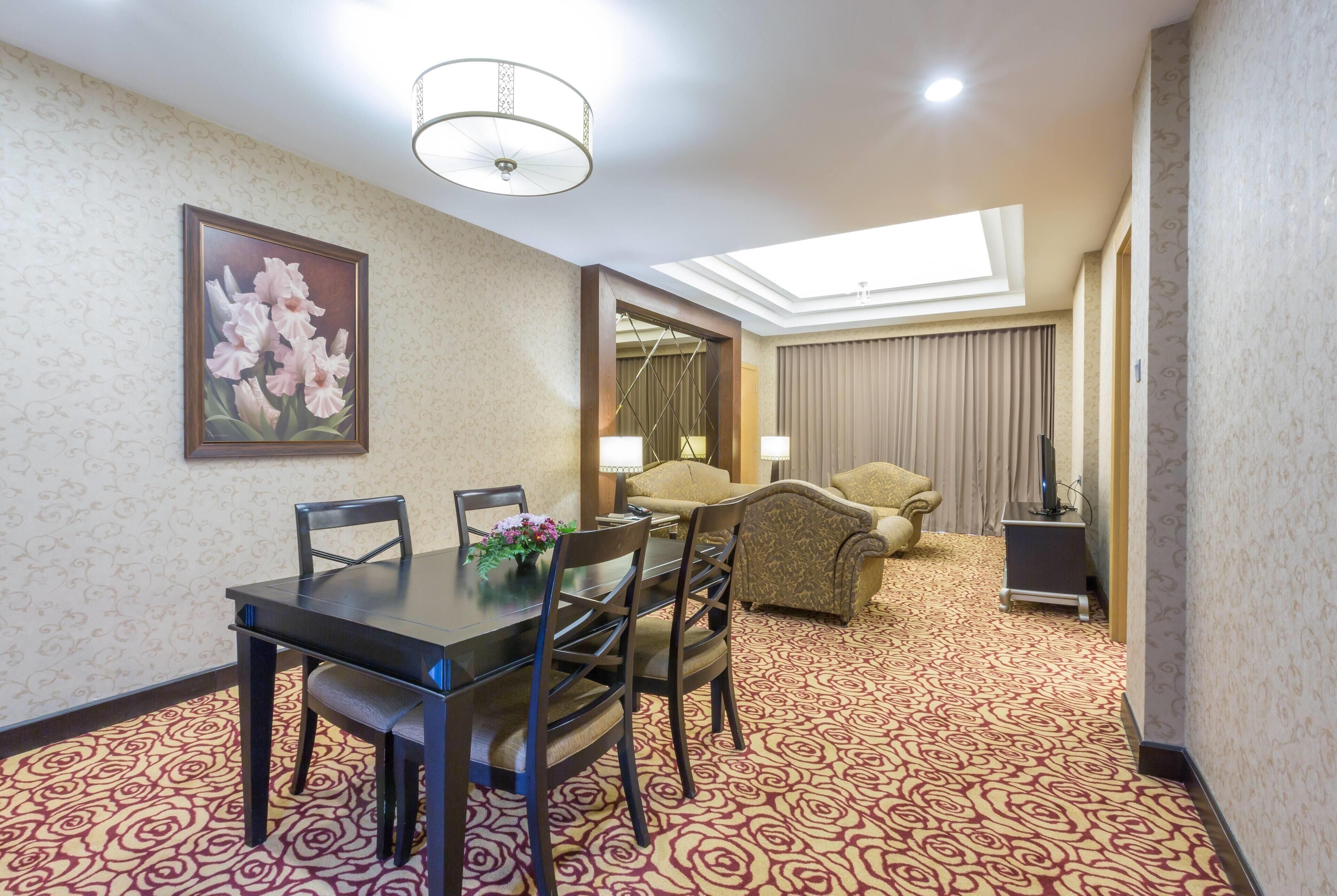 A hotel room with a dining area featuring a table and chairs, a sofa, a television, and a large window with curtains.