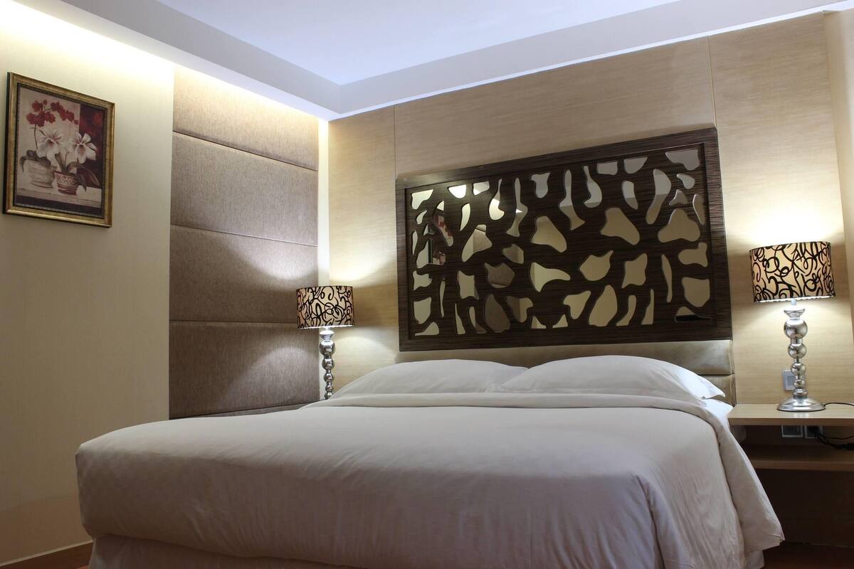 Habitación de hotel con una cama grande, dos lámparas de noche, un cabecero decorativo y un cuadro en la pared.
