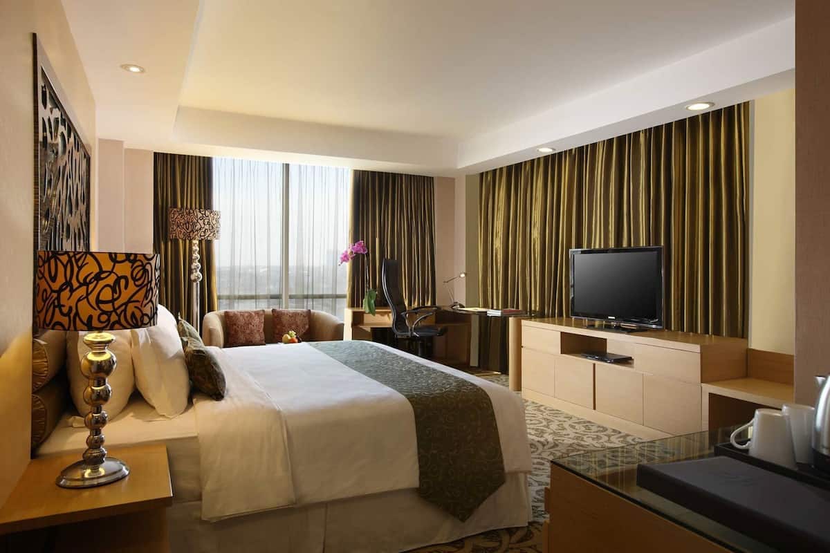 Habitación de hotel con cama, escritorio con televisor, silla, lámpara y ventana con cortinas.