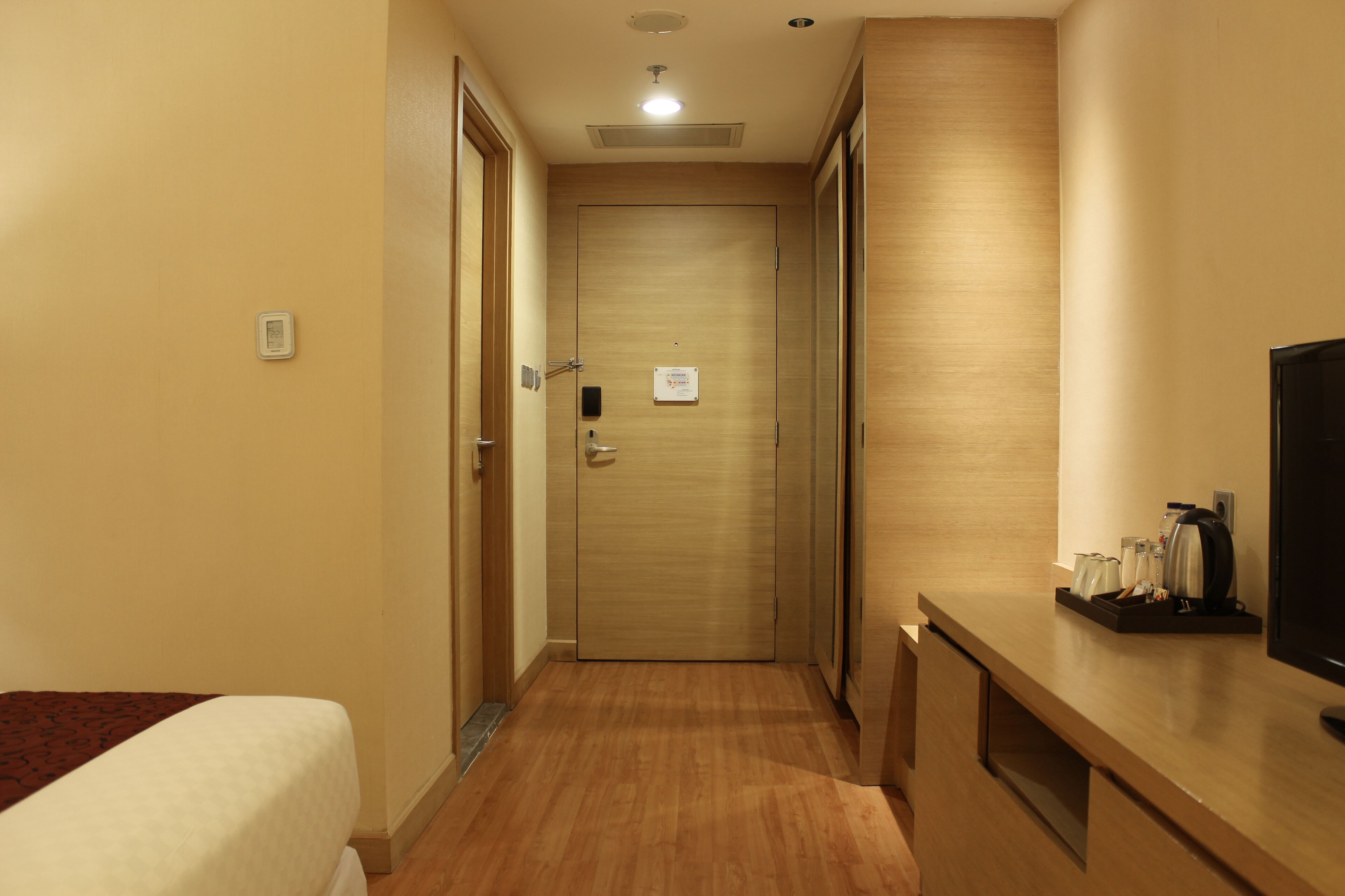 A hotel corridor with a wooden floor, a bed on the left, a desk with a television and coffee maker on the right, and a door at the end.