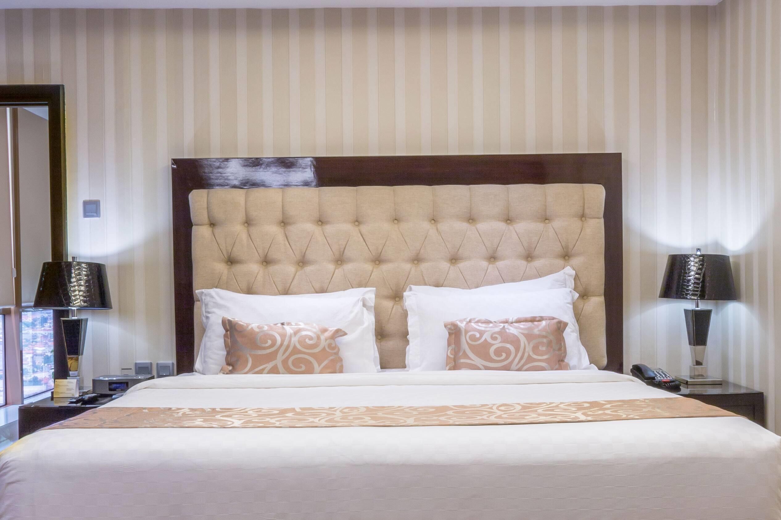 A hotel room with a tufted headboard, a large bed with white and patterned pillows, two bedside tables with lamps, and a view through the window.