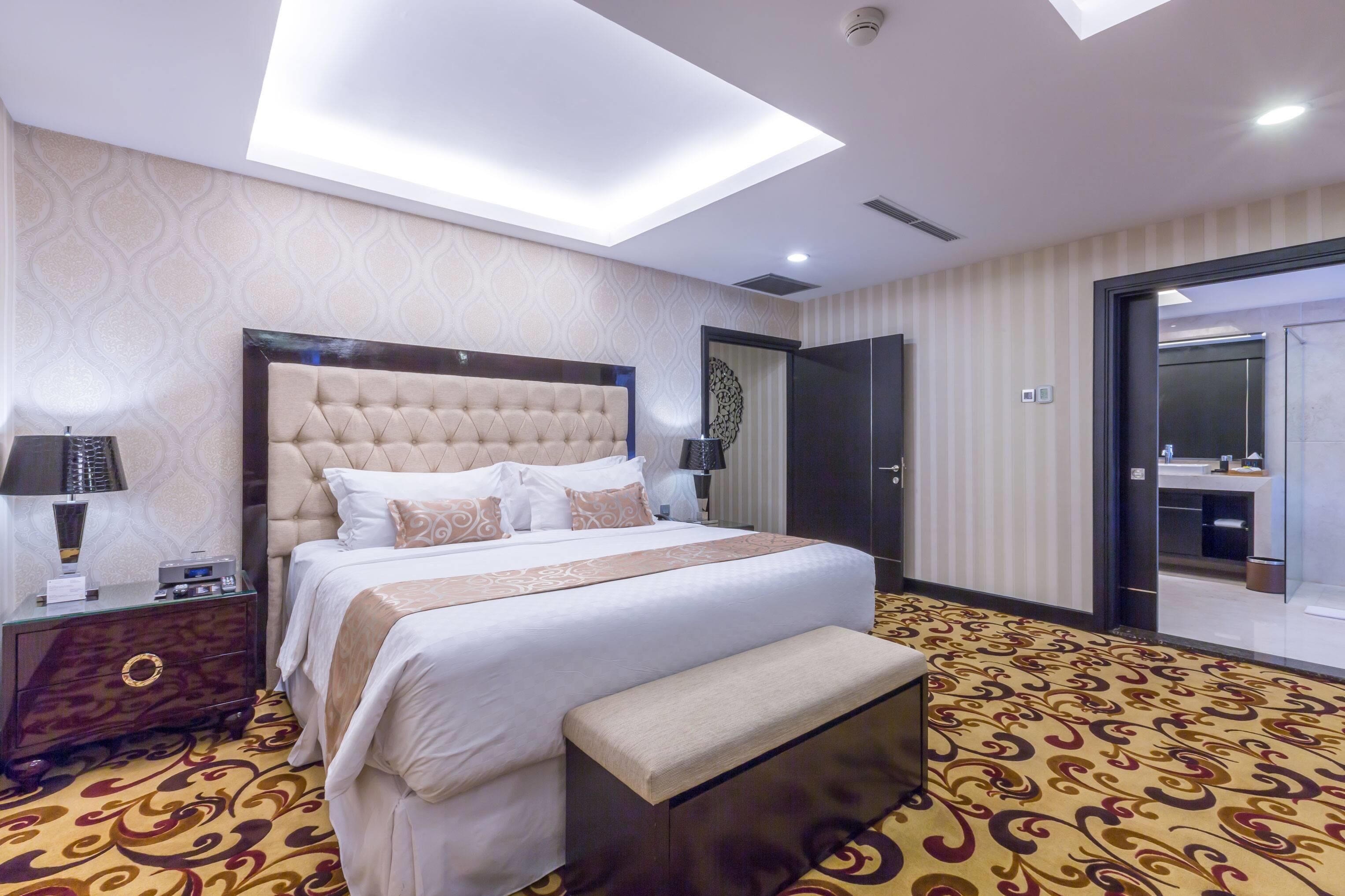 A hotel room with a large bed, a bench, a nightstand, a lamp, and a ceiling light.