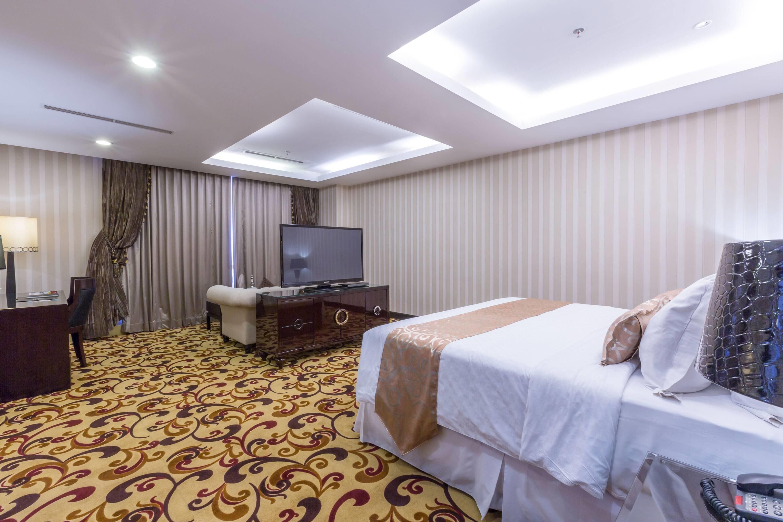 A hotel room with a large bed, a television, a desk with a lamp, and a patterned carpet.