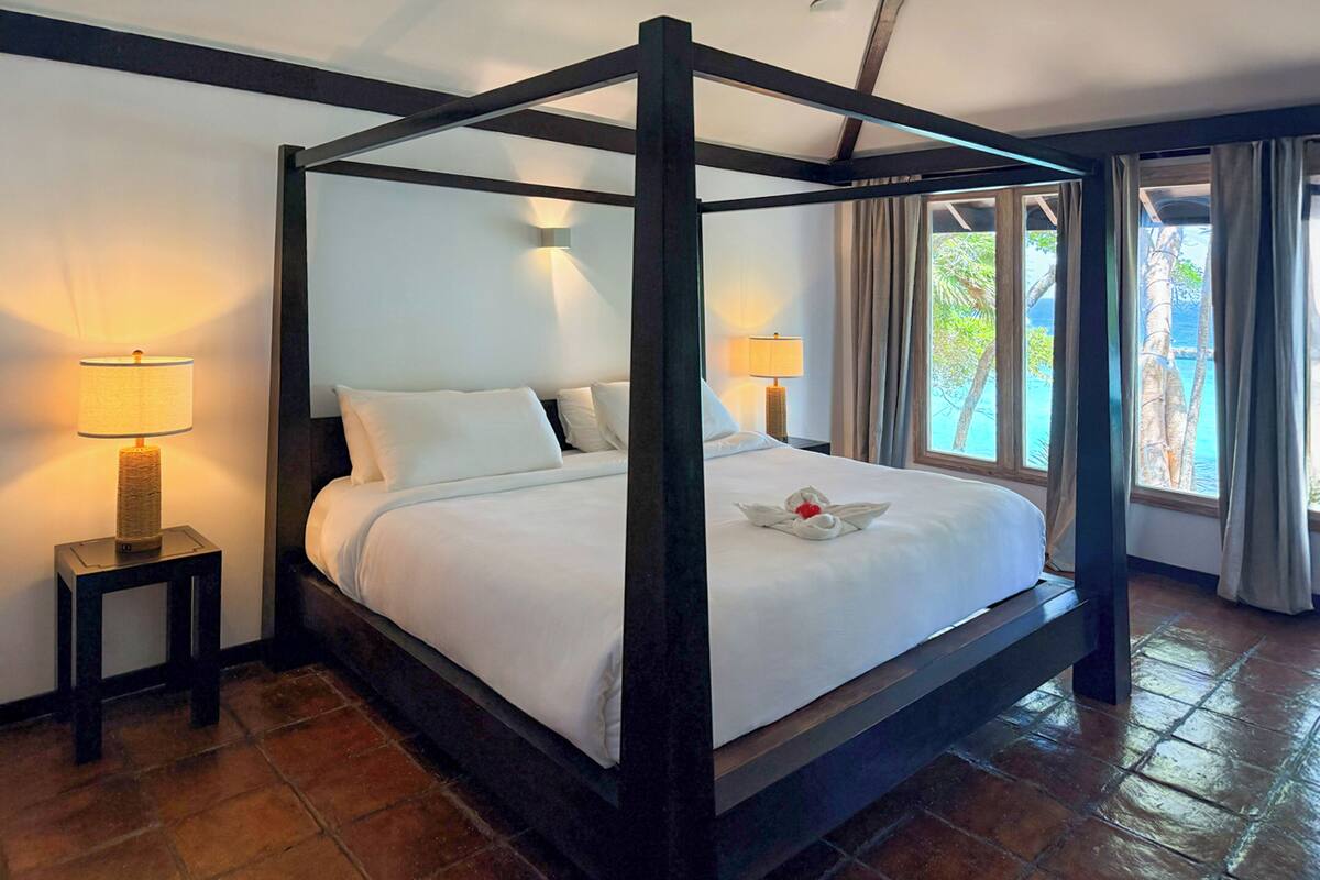 A four-poster bed with white linens, two bedside lamps, and a view of the outdoors through large windows.