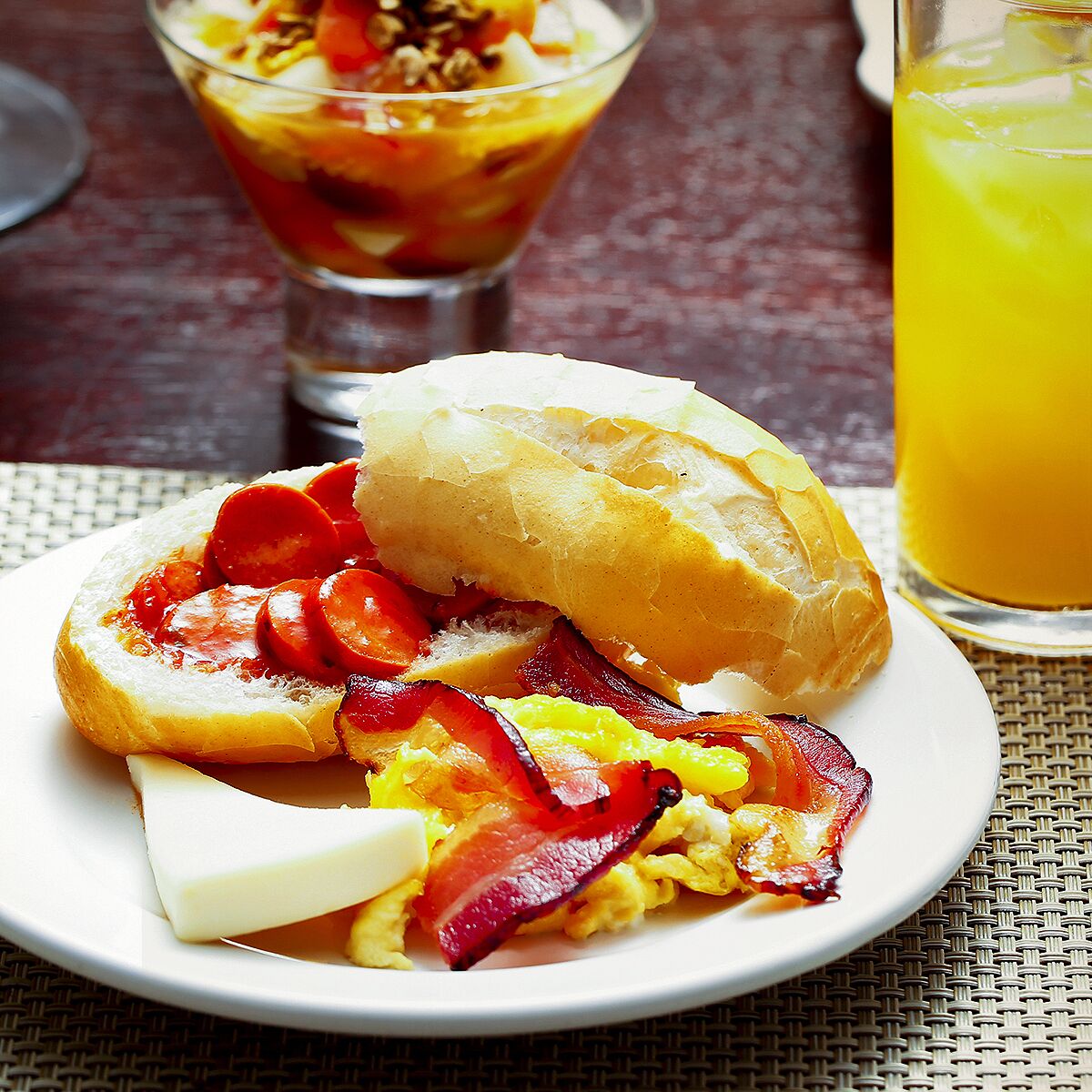 A breakfast plate with eggs, bacon, and tomato slices on a bun, a glass of orange juice, and a bowl of fruit salad in the background.