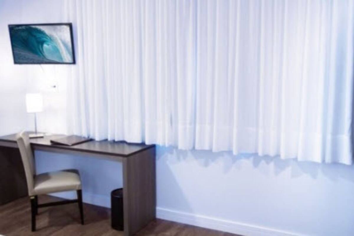 A hotel room with a desk, chair, and a large window with white curtains.