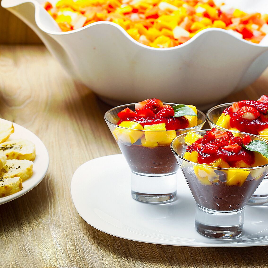 Three glasses of dessert with fruit on top, a white bowl of diced fruit in the background, and a plate with more fruit pieces on a wooden table.