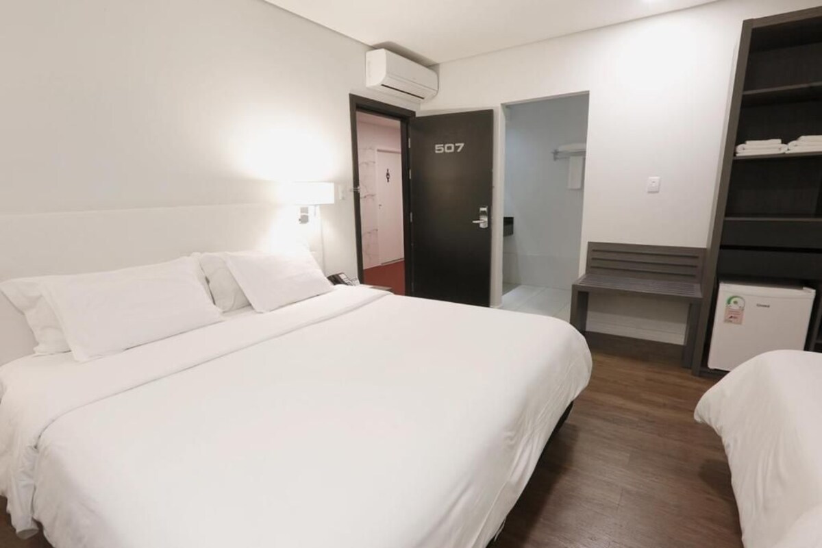 A hotel room with a bed, a desk, a wardrobe, and an air conditioning unit.