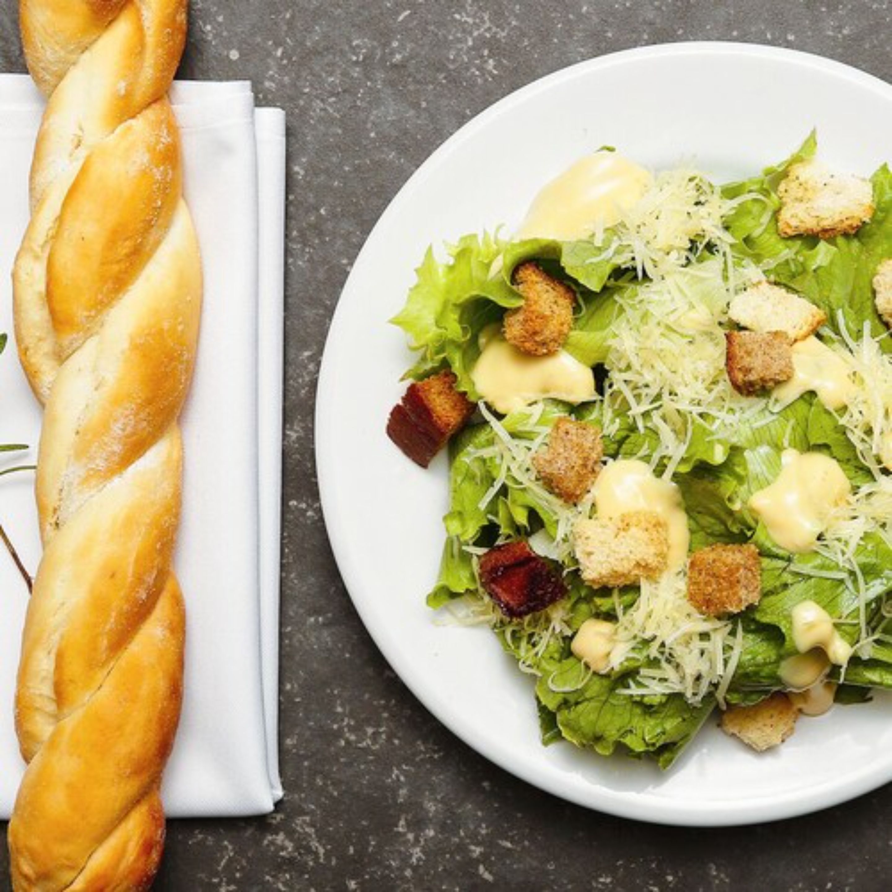 Una ensalada con crutones y queso, y un pan trenzado sobre una superficie oscura.