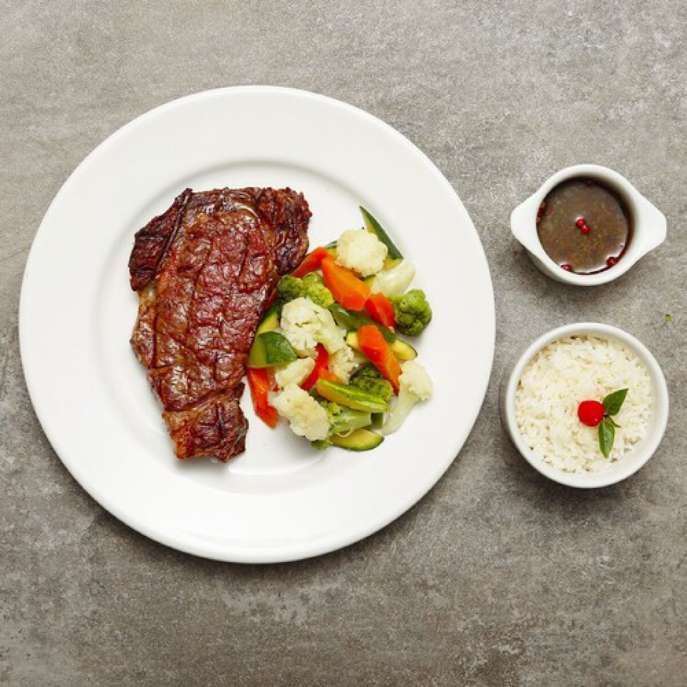 A plate with a steak, mixed vegetables, and two small bowls of sauce.