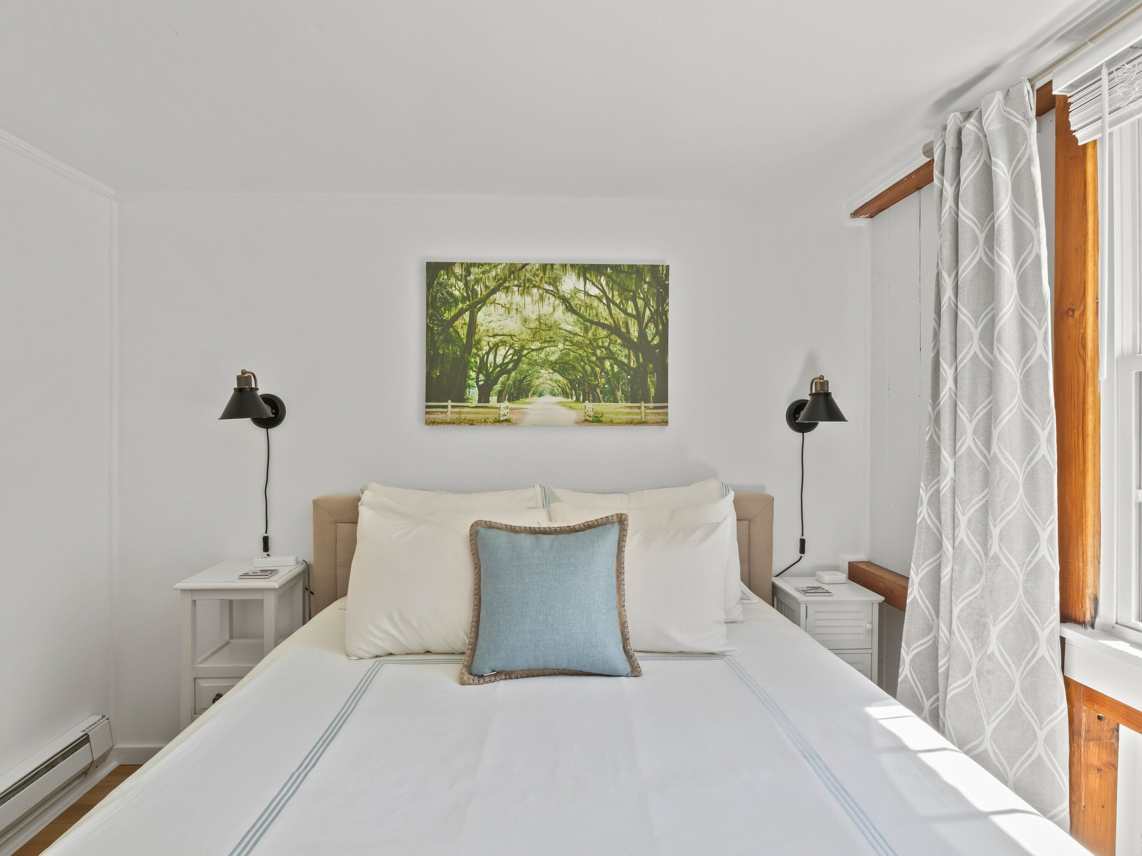 A bedroom with a large bed, two bedside tables, a nightstand, a wall-mounted lamp, a window with curtains, and a picture of a forest.