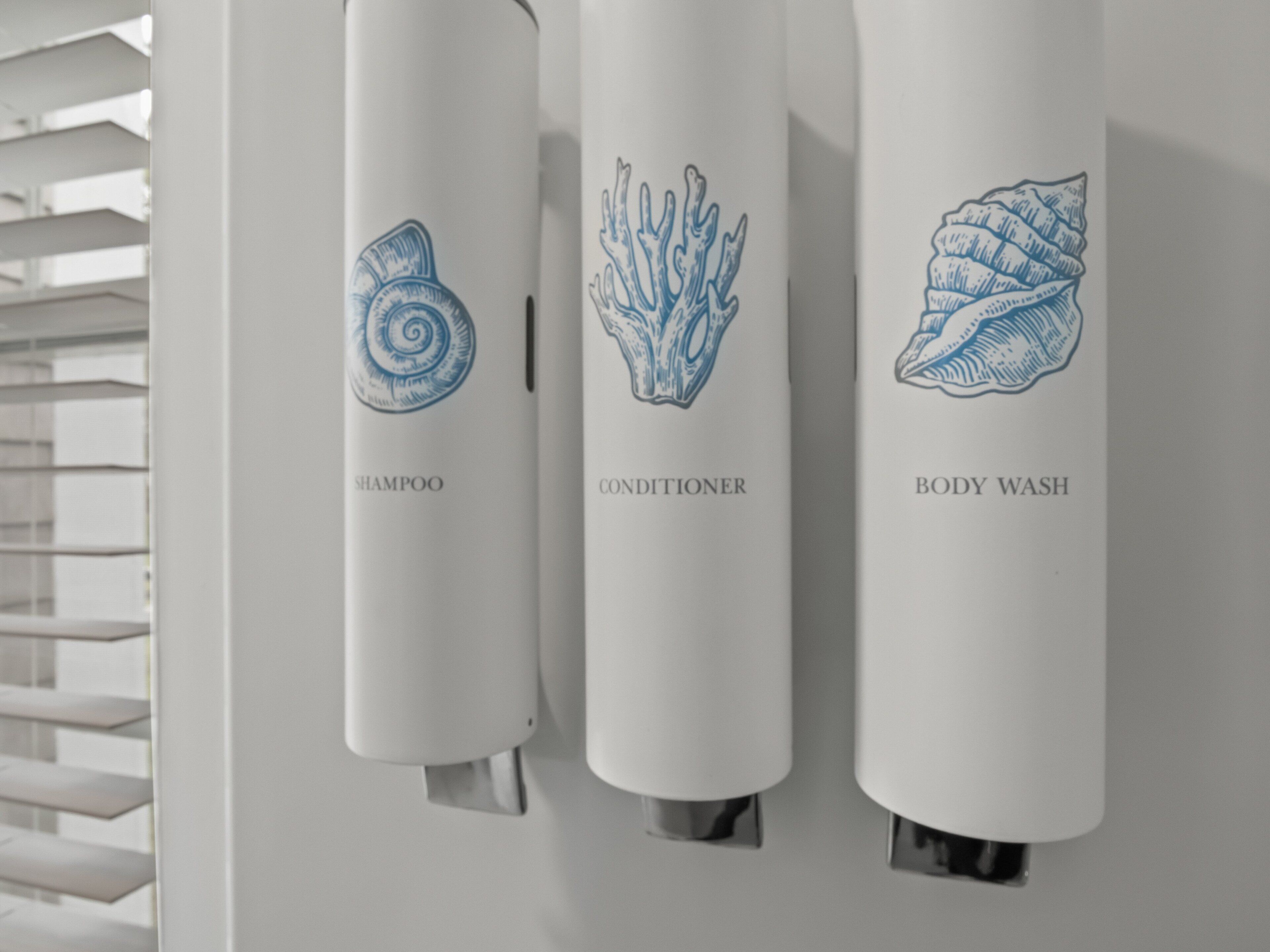 Trois distributeurs étiquetés « SHAMPOO », « APR ÈS-SHAMPOO » et « GEL DOUCHE », et illustrés de motifs représentant des coquillages.