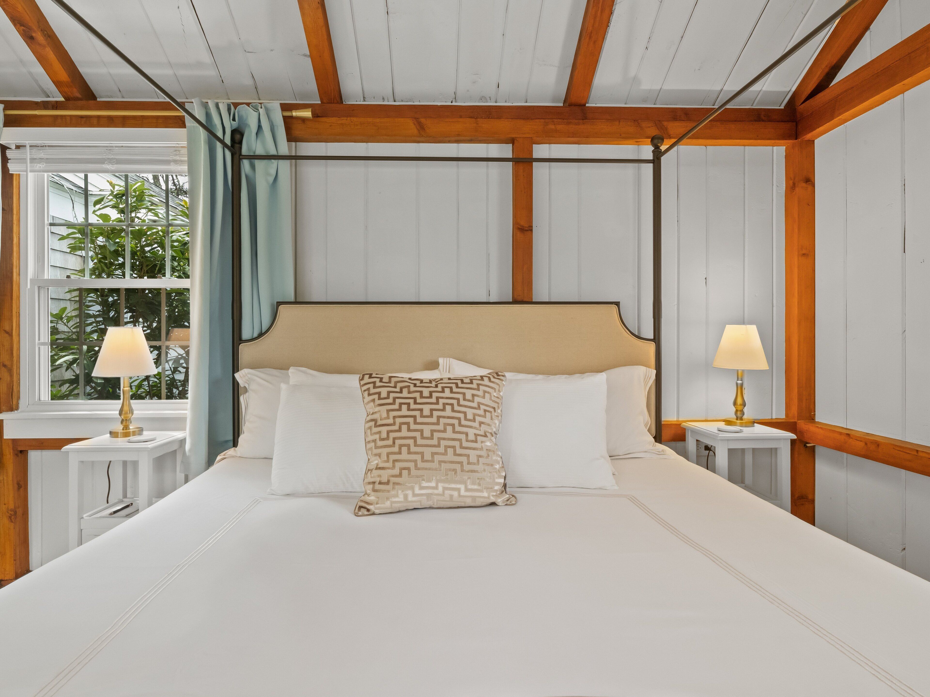 A bedroom with a large bed, two bedside tables with lamps, wooden beams on the ceiling, and a window with curtains.