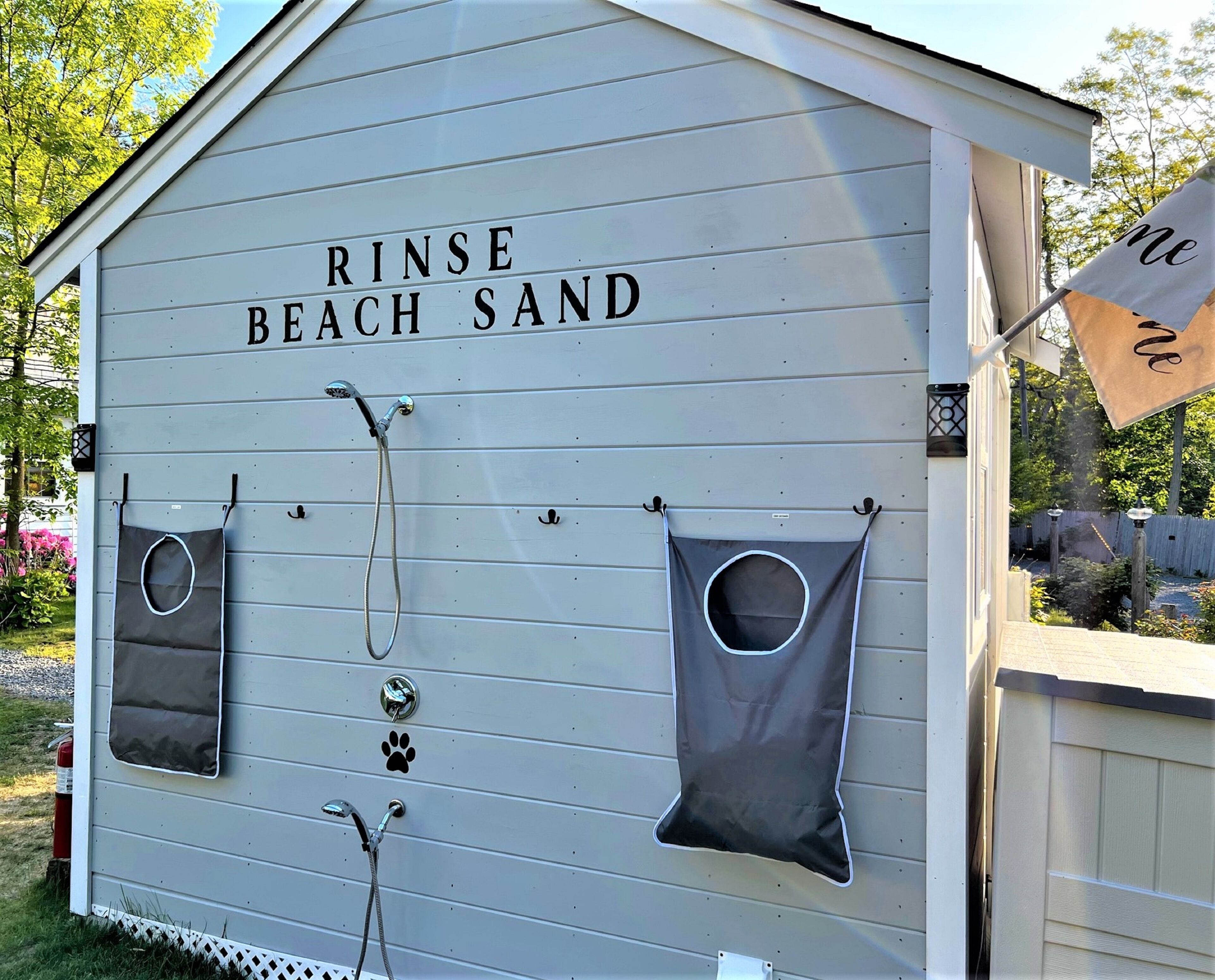 A small building labeled "RINSE BEACH SAND" with two showerheads and two small bags hanging from the wall.