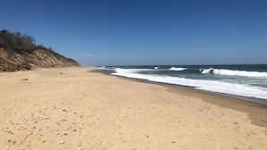 Uma praia arenosa com ondas, céu claro e um penhasco rochoso.