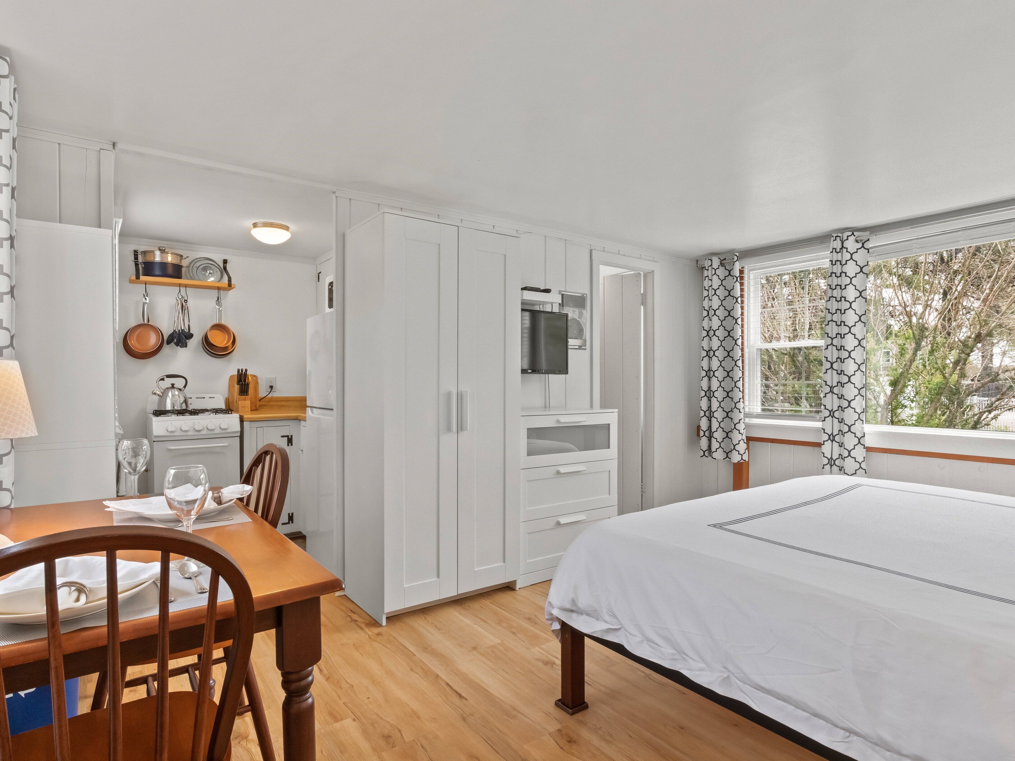 A bedroom with a bed, a wooden dining table with chairs, a kitchen area with cabinets and appliances, and a window with patterned curtains.