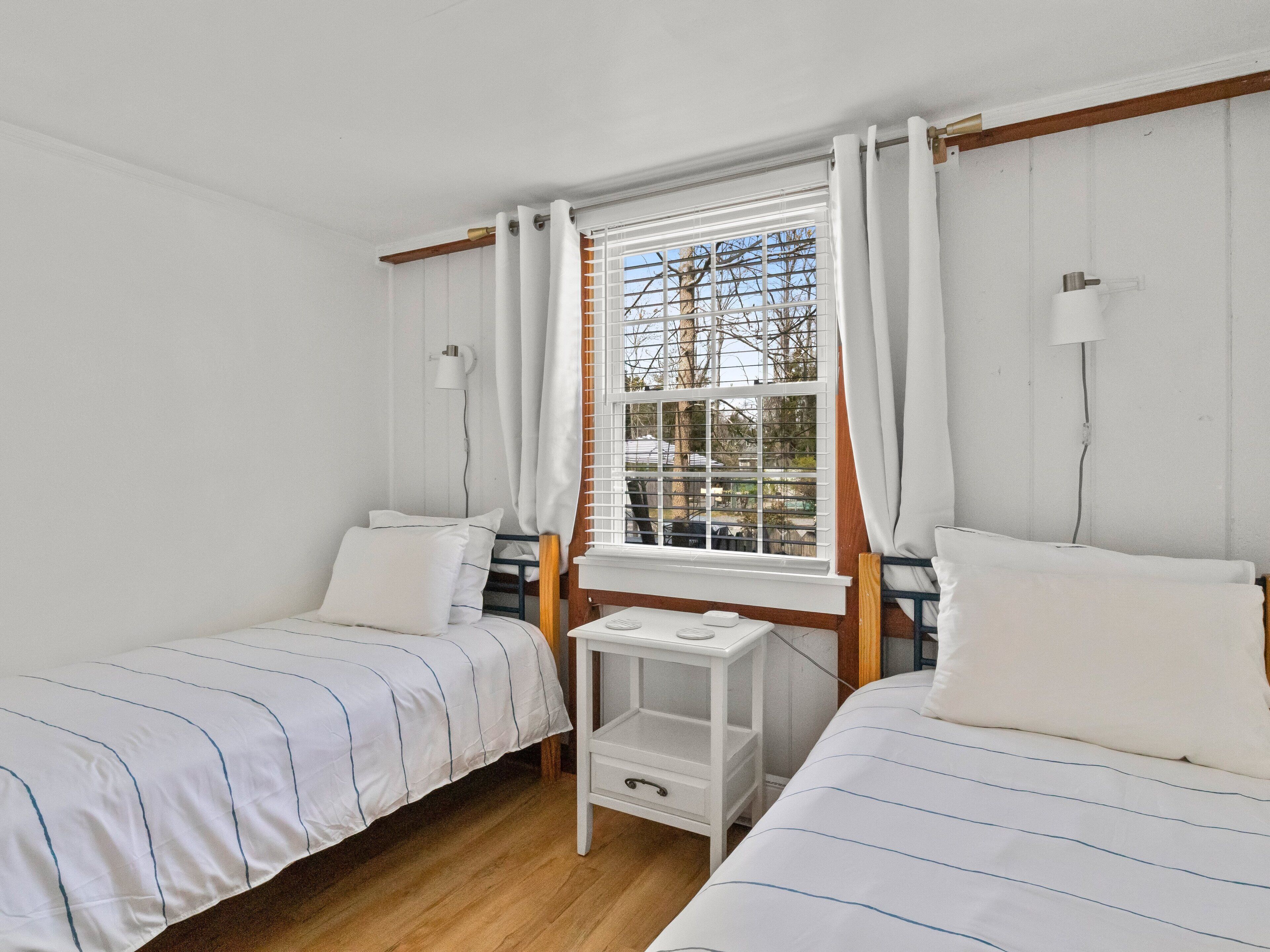 A small, tidy room with two single beds, a small table, and a window with blinds.