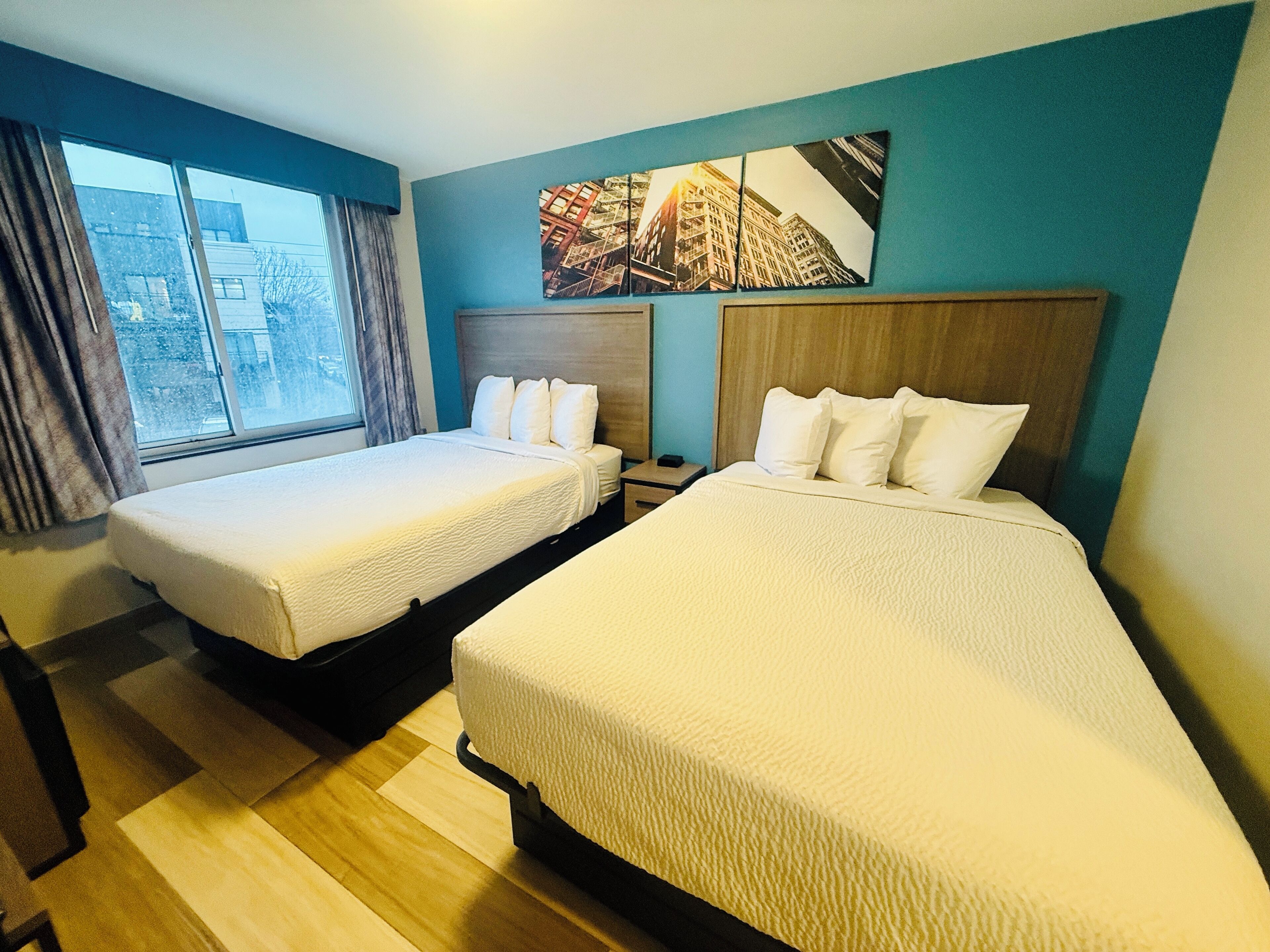 Une chambre d’hôtel avec deux lits, une fenêtre avec des rideaux, une table de chevet et un tableau représentant un paysage urbain accroché au mur.