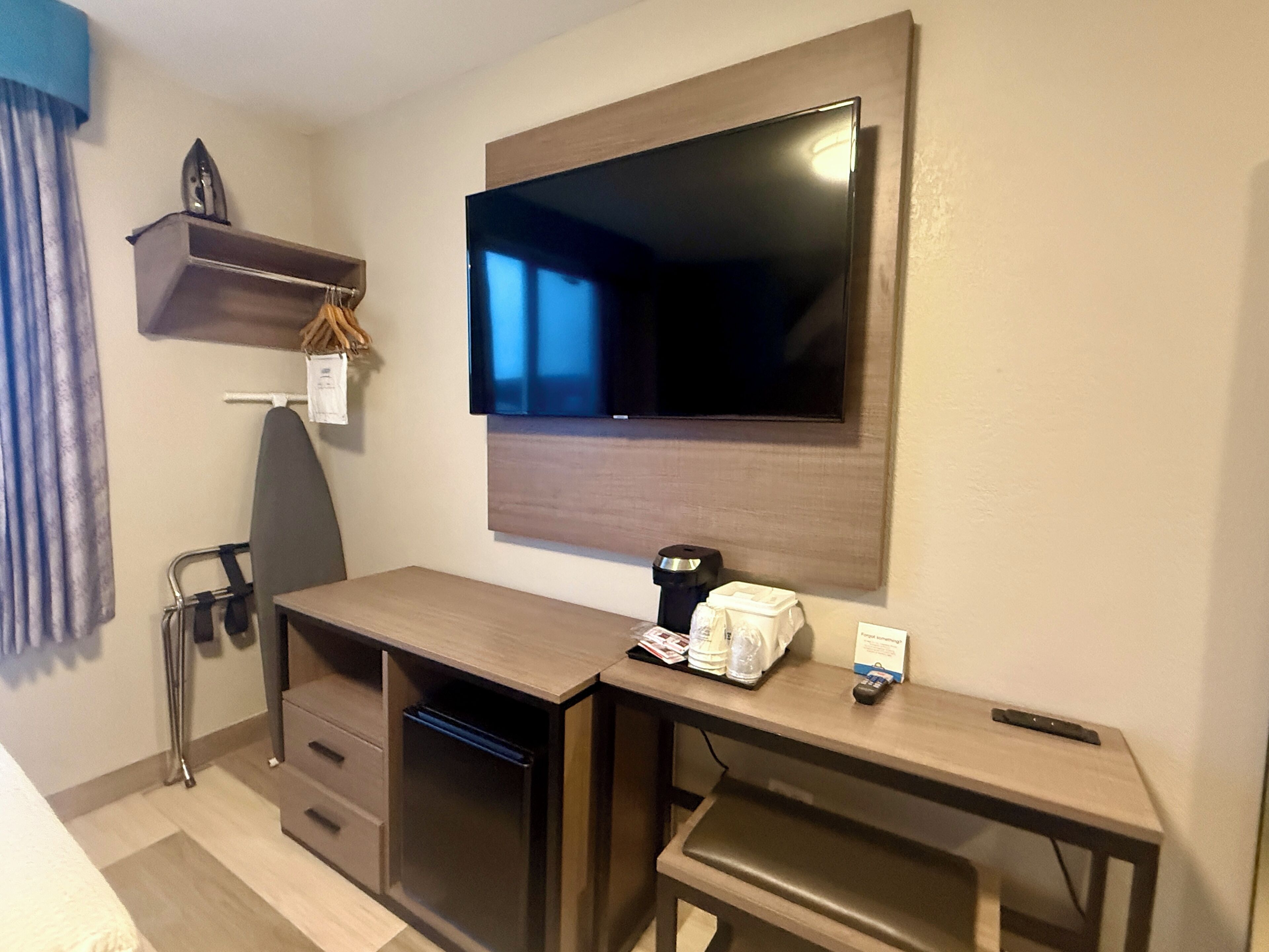 A compact hotel room with a wooden desk, a flat-screen TV mounted on the wall, a small shelf with a decorative item, and a chair.