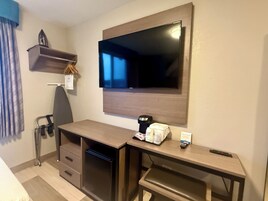 A compact hotel room with a wooden desk, a flat-screen TV mounted on the wall, a small shelf with a decorative item, and a chair.