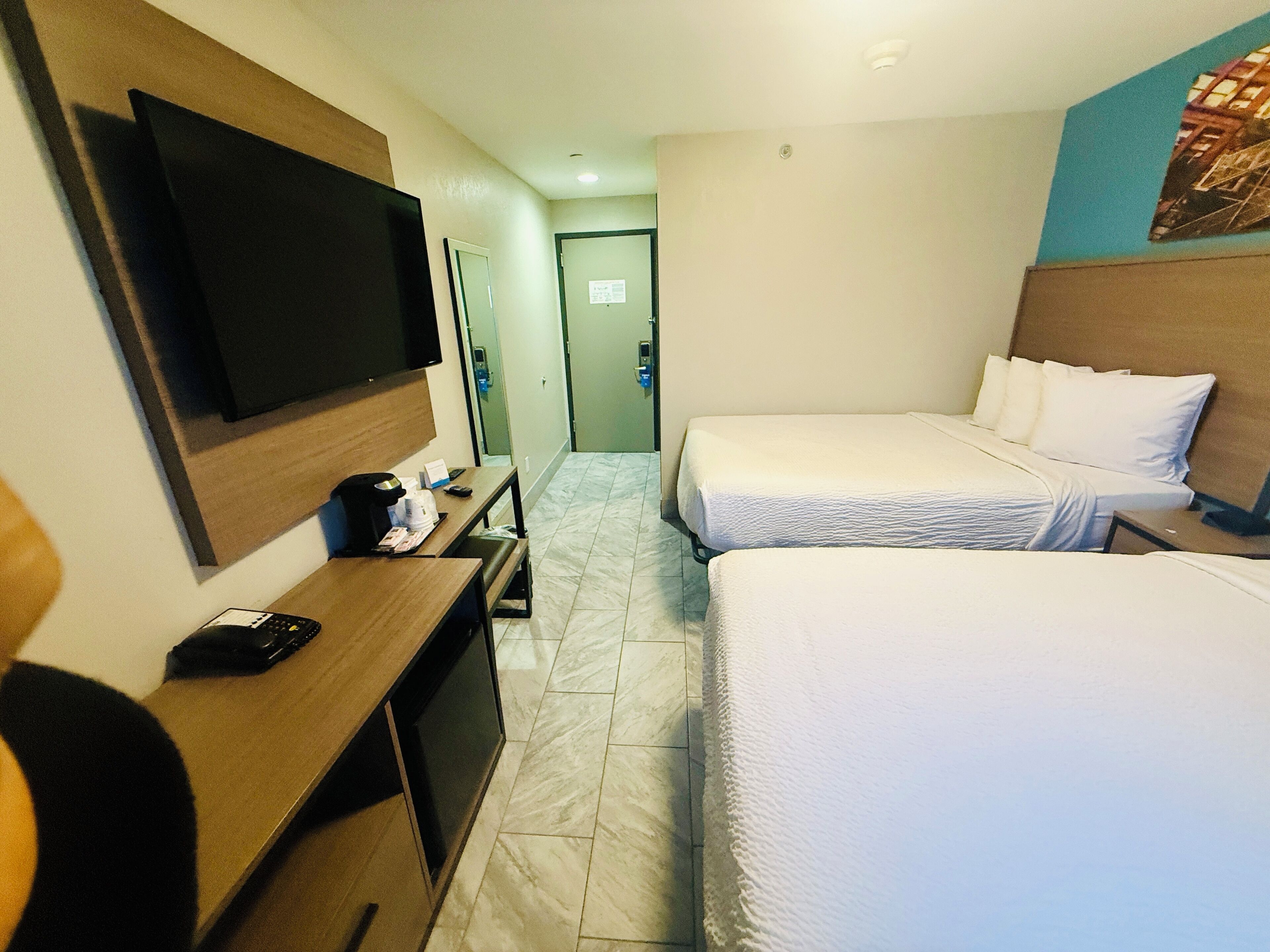 Habitación de hotel con dos camas, un escritorio, televisor de pantalla plana, cafetera y una puerta que da a un pasillo.