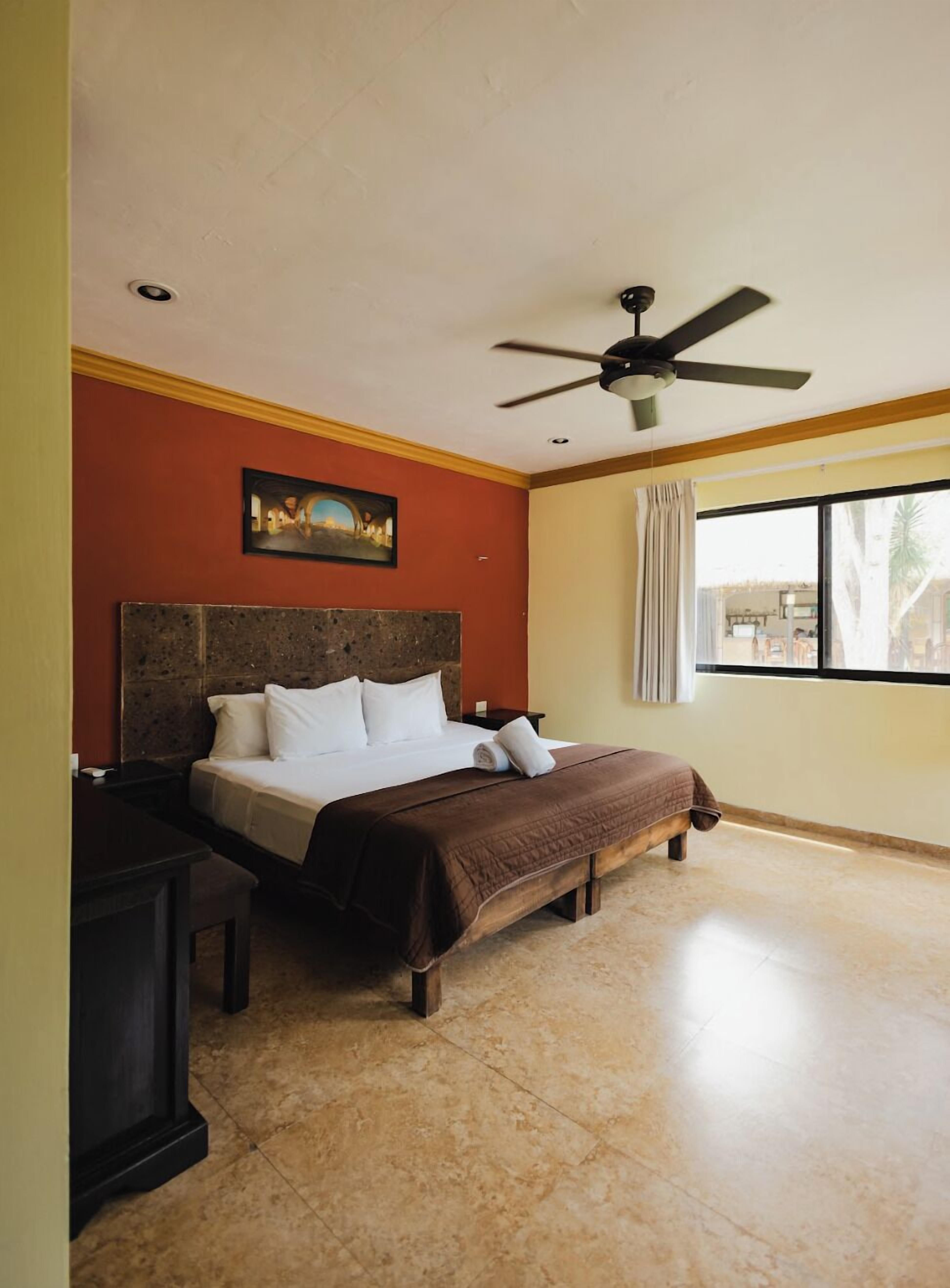 Habitación de hotel con cama, ventilador de techo, un cuadro en la pared y una ventana con cortinas.
