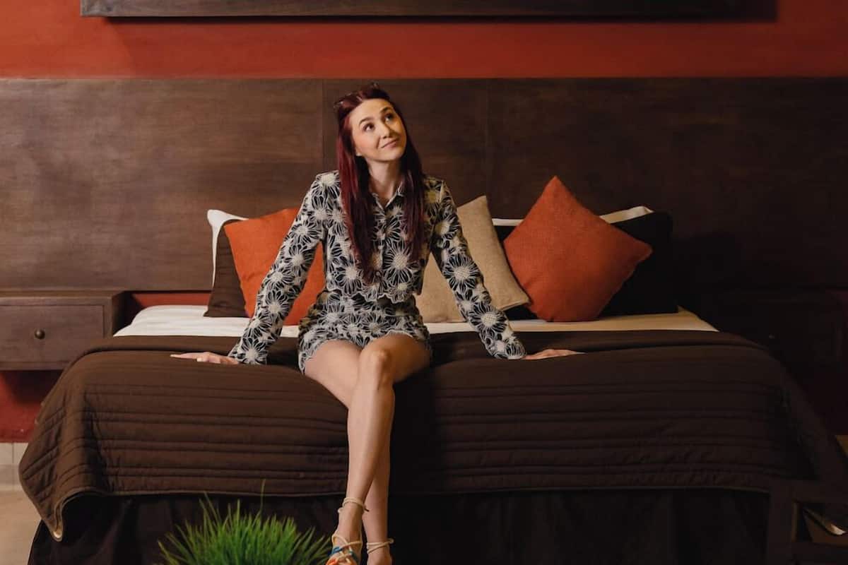 A woman sitting on a bed in a room with a ceiling fan, a shelf with plants, and a potted plant on the floor.
