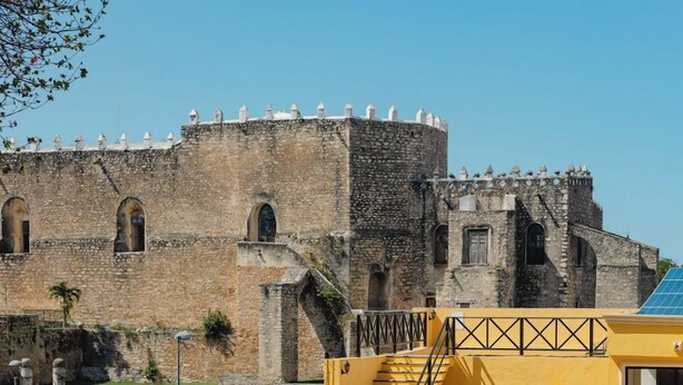 Un edificio moderno amarillo con piscina, un muro de piedra y un edificio histórico de piedra al fondo.
