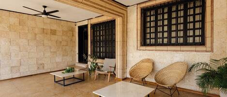 Una habitación con suelo de baldosas, ventilador de techo, una mesa, dos sillas de mimbre y una ventana con barrotes.