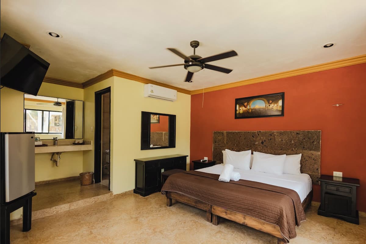 A hotel room with a bed, a ceiling fan, a TV, a sink, and a painting on the wall.