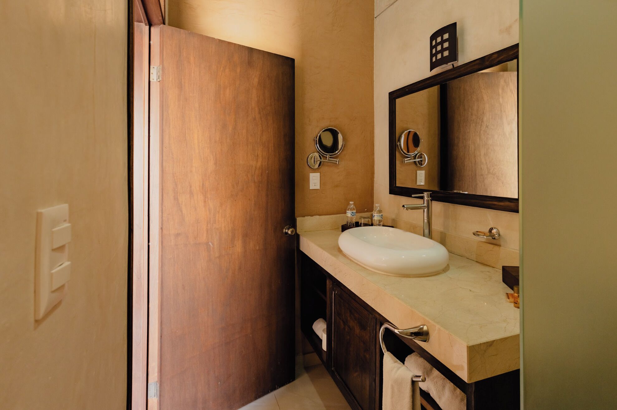 Un baño con puerta de madera, un lavabo blanco, un espejo y un perchero para toallas.