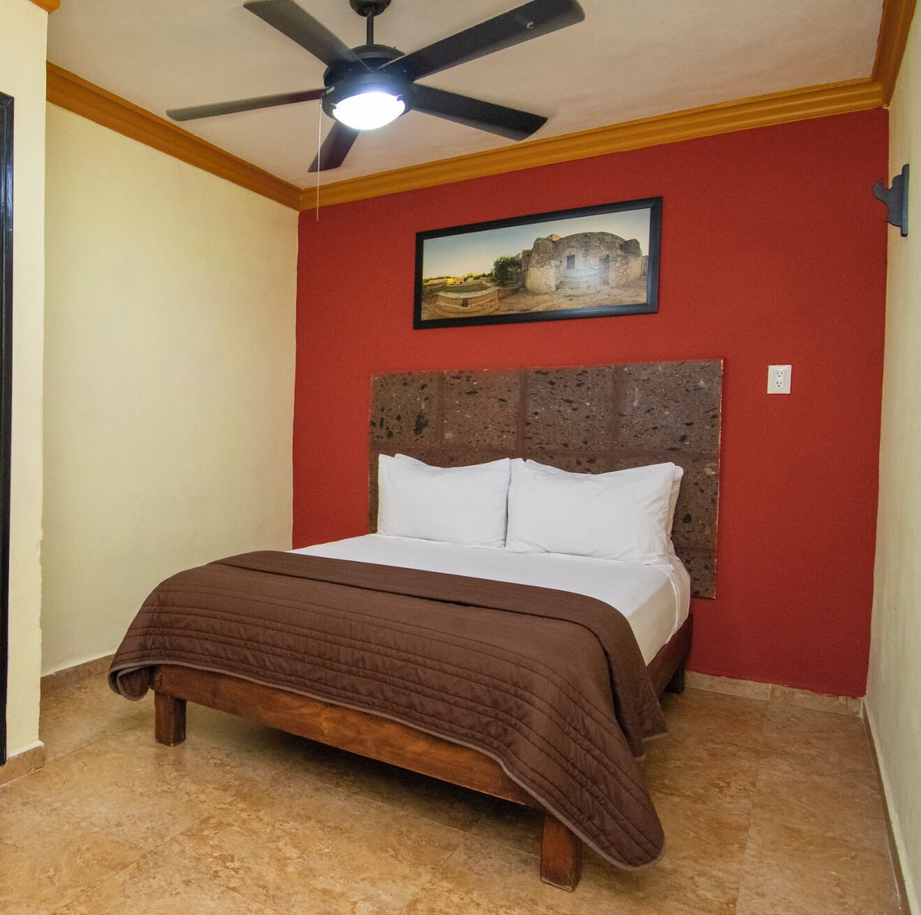 A bedroom with a bed, a ceiling fan, a red wall, and a framed picture.