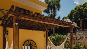 Un espacio de descanso techado al aire libre con dos sillones reclinables y una hamaca, un pérgola de madera y un edificio amarillo al fondo.