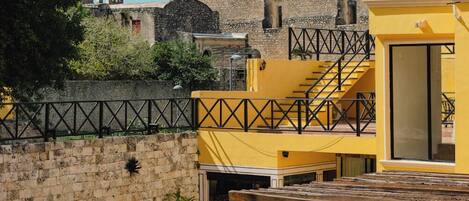 Un edificio moderno amarillo con piscina, terraza de madera y un muro de piedra con una estructura al fondo que recuerda a una fortaleza.