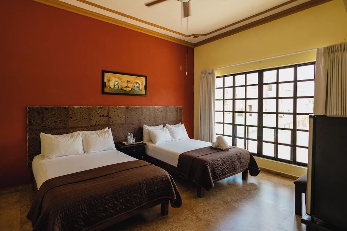 A hotel room with two beds, a ceiling fan, a large window with curtains, and a painting on the wall.