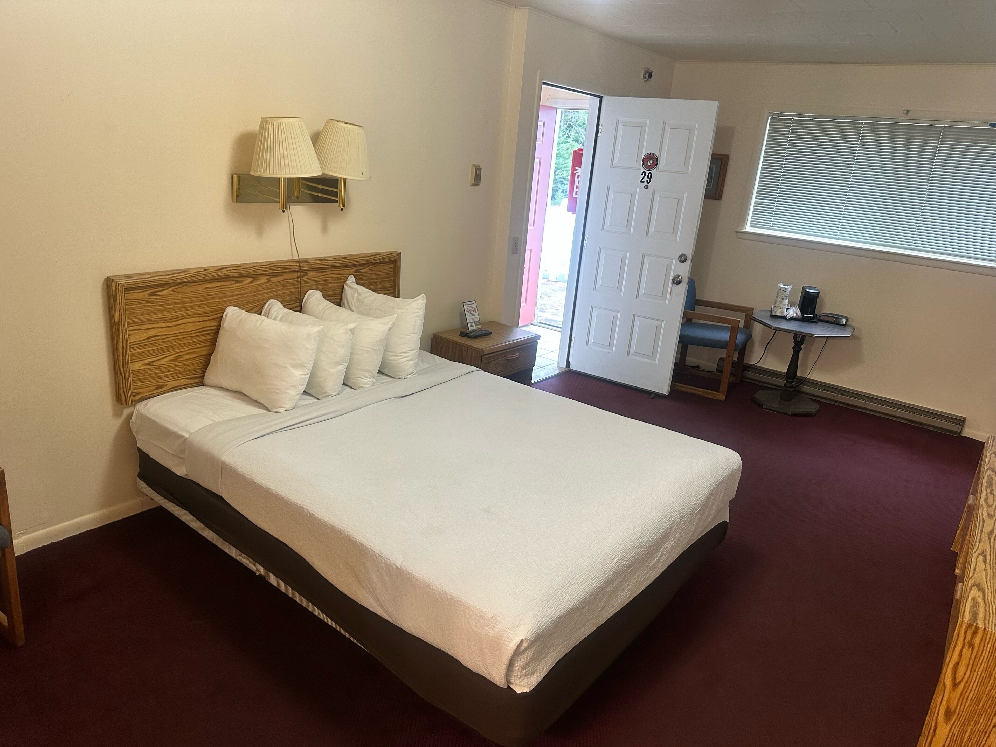 Una camera d'albergo con un letto grande, una testiera in legno, due lampade ai comodini, un tavolino con una sedia, una porta e una finestra con persiane.
