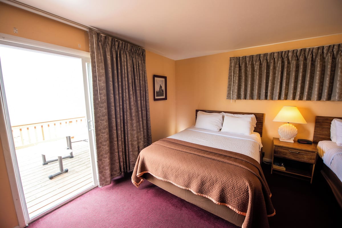 Un cuarto de hotel con una cama, una mesita de noche con lámpara, una ventana con cortinas y vistas al área exterior.