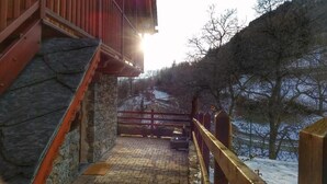 Eine Steintreppe führt zu einem Holzbalkon mit Metallgeländer, der auf eine verschneite Landschaft blickt.