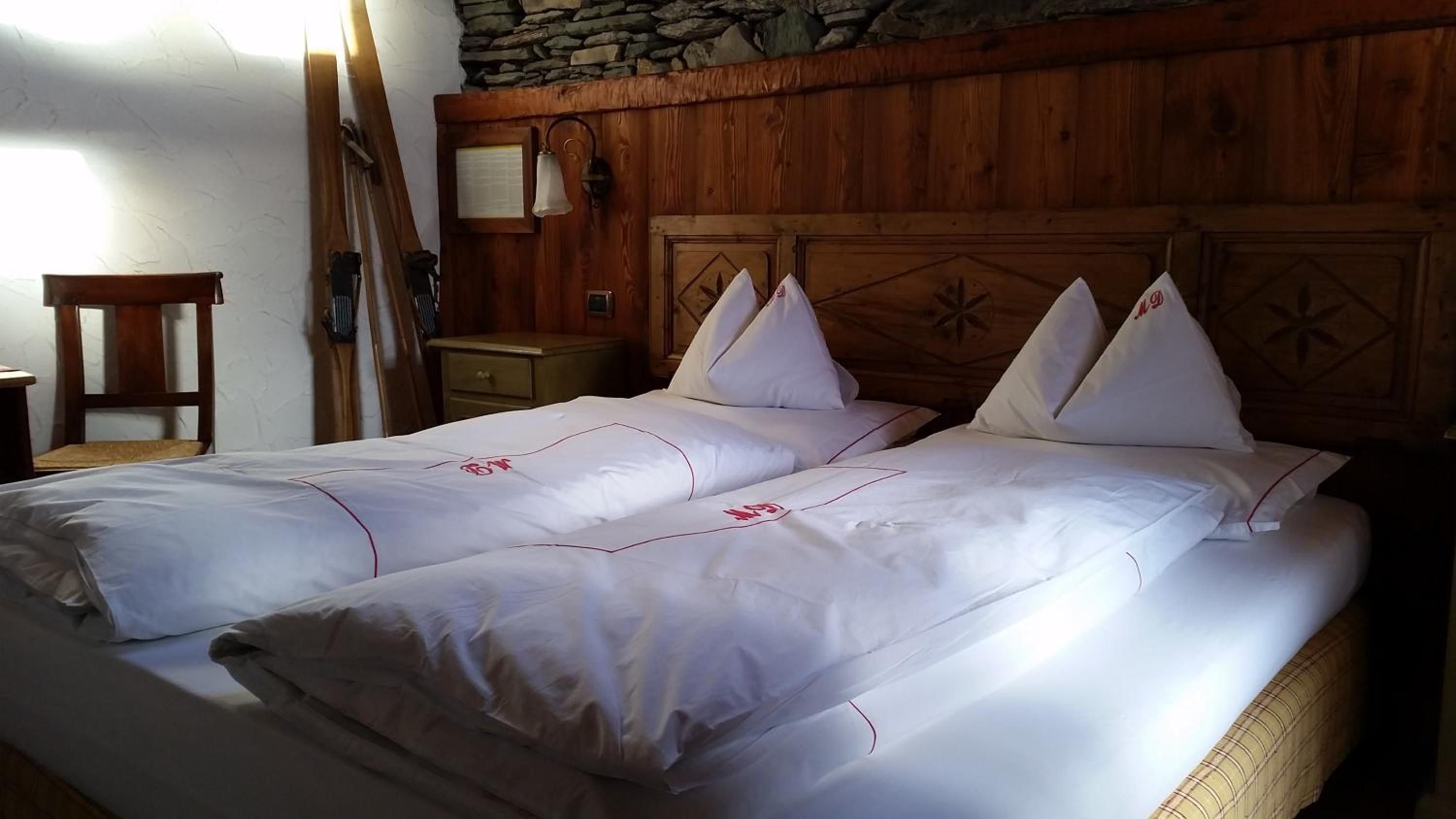 A double bed with white bedding and red accents, a wooden headboard with a geometric pattern, a nightstand with a lamp, and a stone wall.