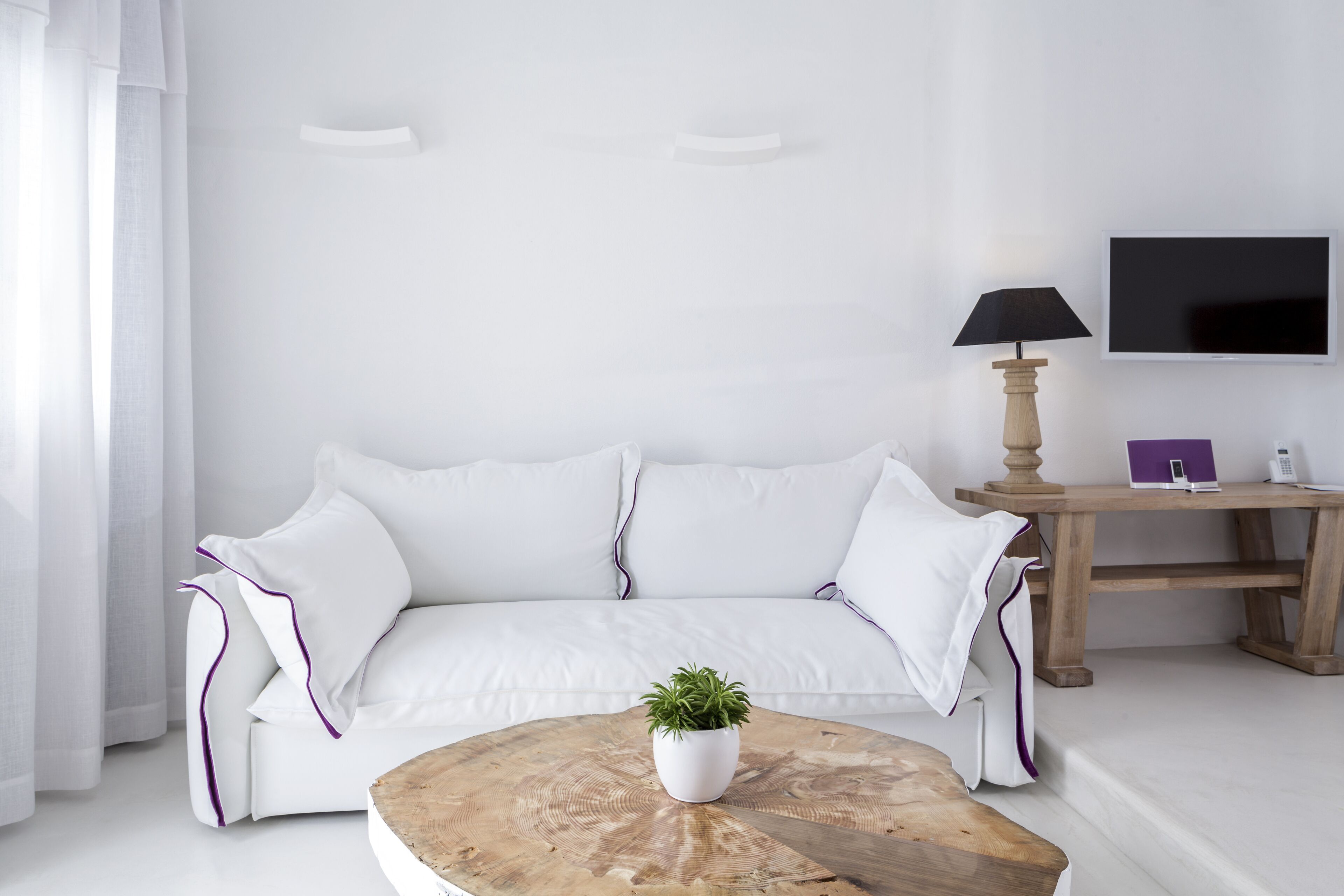 A modern living room with a white sofa, a round coffee table with a plant, a wooden side table with a lamp, and a flat-screen TV mounted on the wall.
