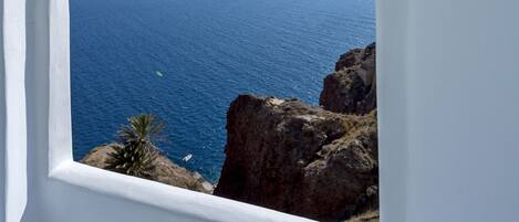A view through an arched window overlooking a coastal landscape with a clear blue sea and rocky terrain.