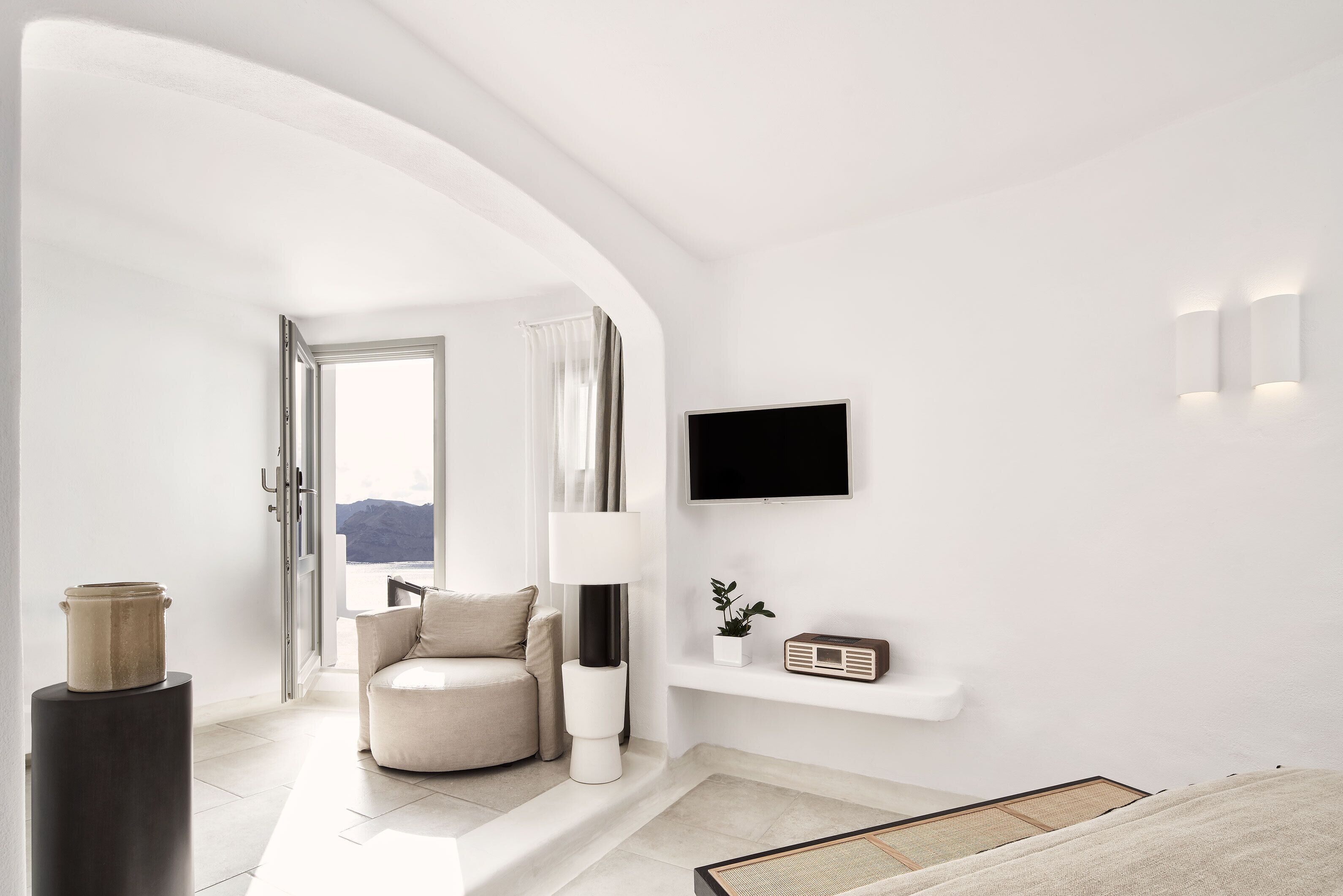 A modern, minimalist living room with a sofa, a TV mounted on the wall, and a small shelf with a plant and a radio.