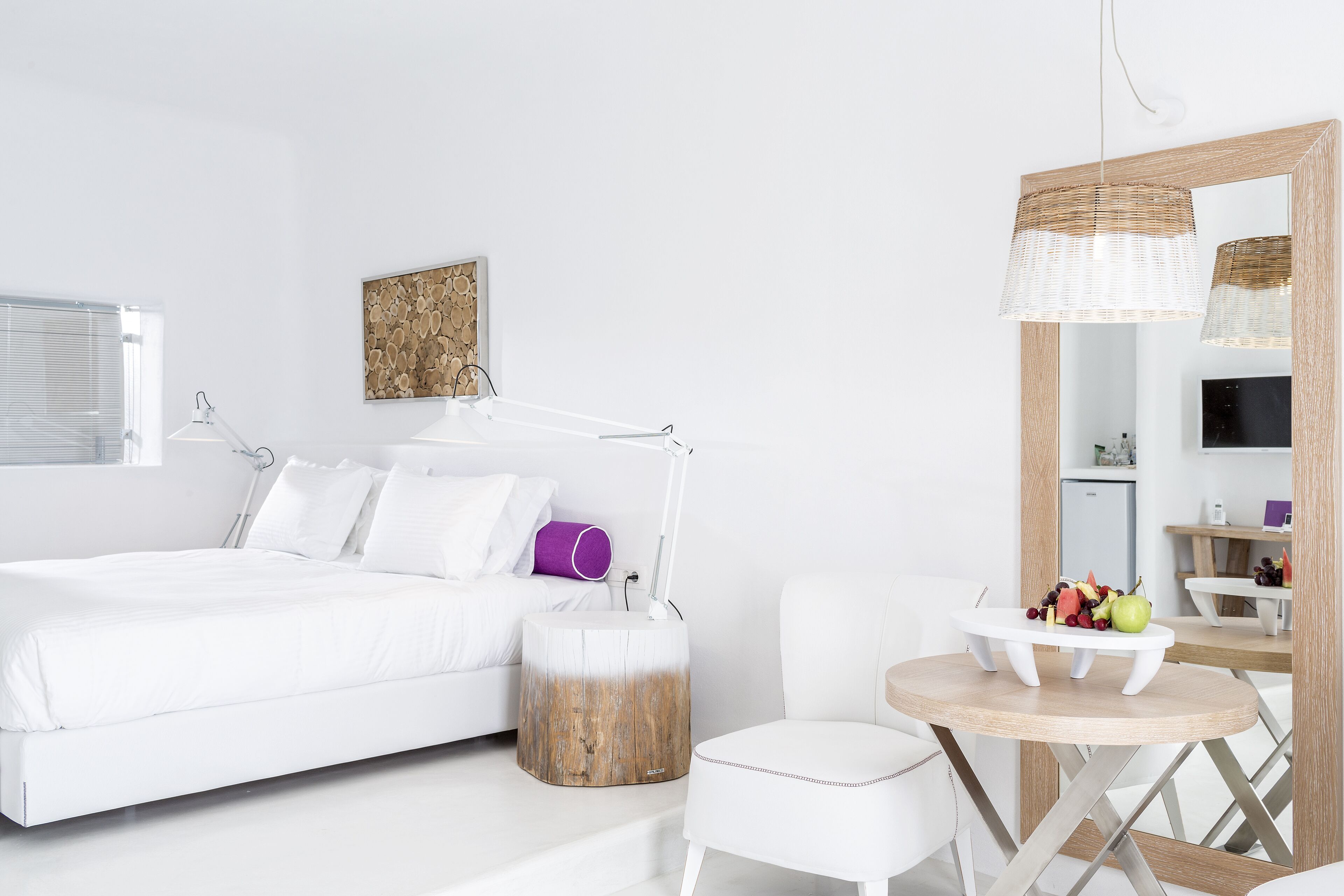 A modern, minimalist bedroom with a white bed, bedside table, and a round wooden table with fruit.