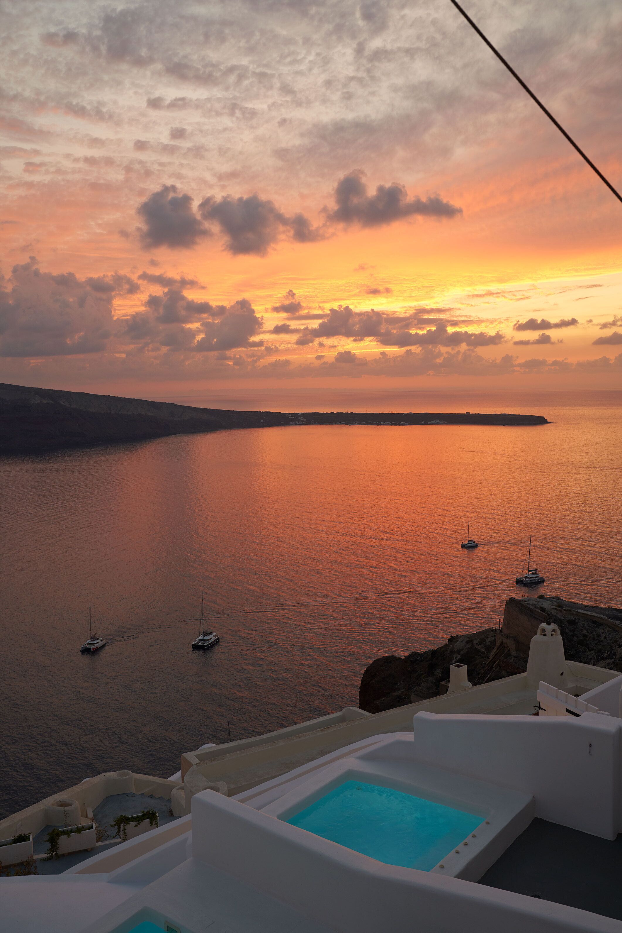 A sunset view over a calm sea with boats, a white pool, and a rocky coastline.