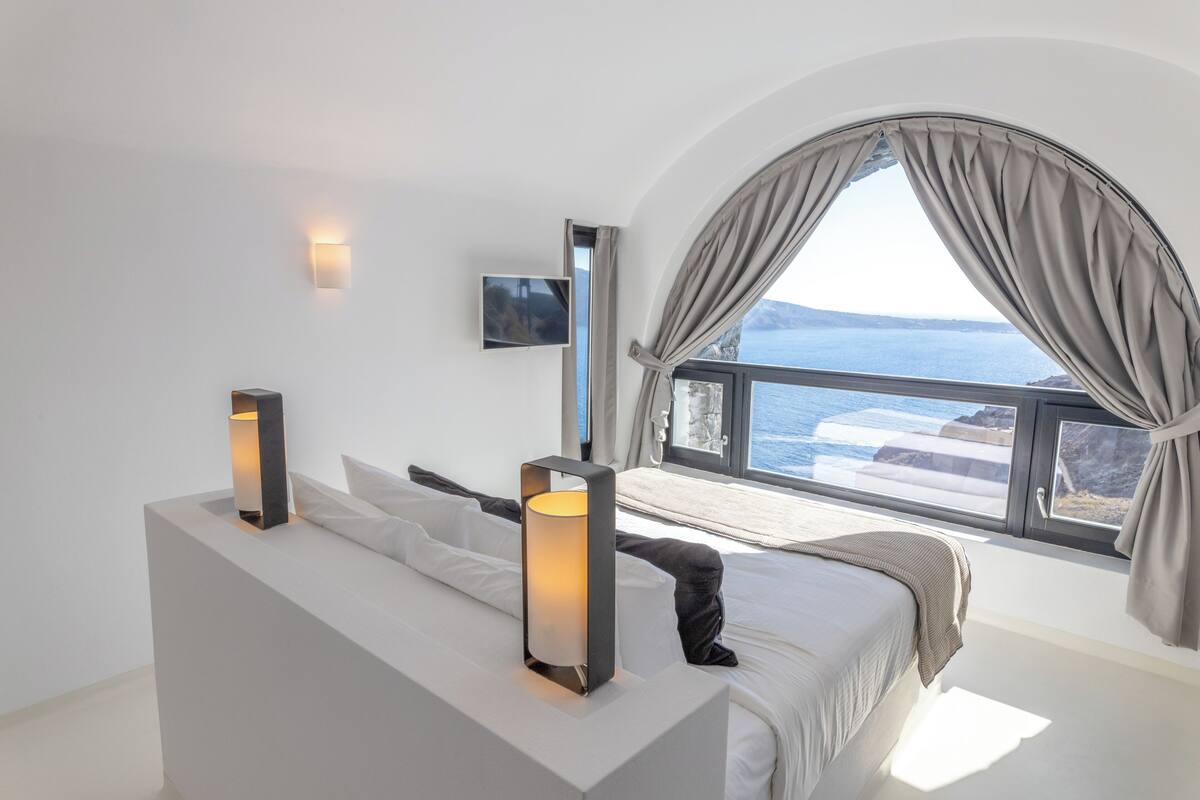 A modern bedroom with a large bed, a bedside table, a TV, and a view of the sea.