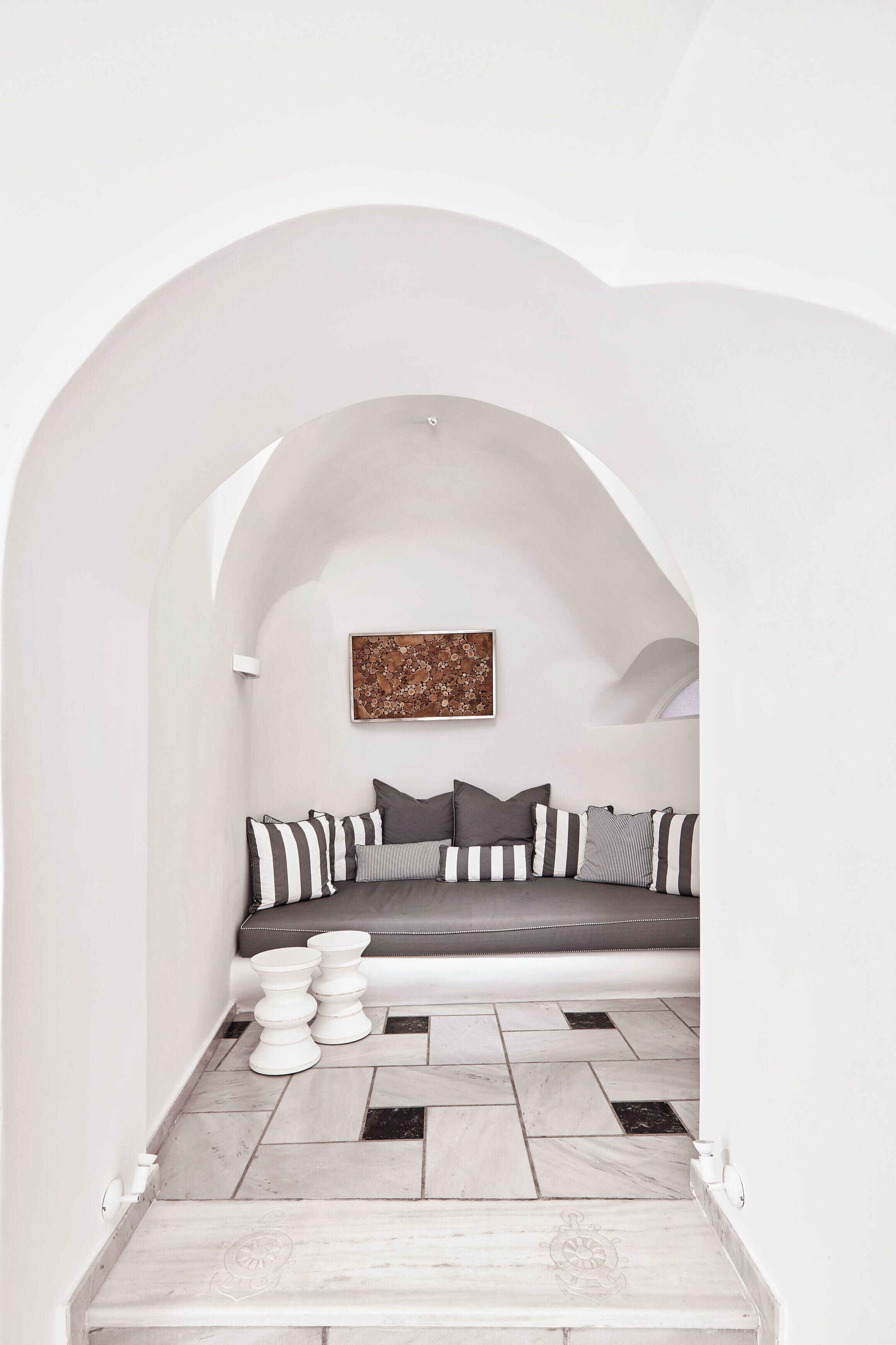 Una habitación minimalista con un arco blanco que da a una zona de estar con un sofá y cojines. Una obra enmarcada se exhibe en la pared sobre el sofá.